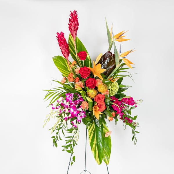 Tall tropical standing spray with orange lilies, red roses, purple orchids, birds of paradise, and large tropical leaves.