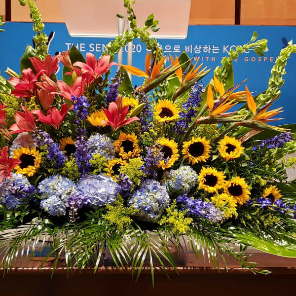 Large stage arrangement with sunflowers, pink lilies, blue hydrangeas, and orange birds of paradise.