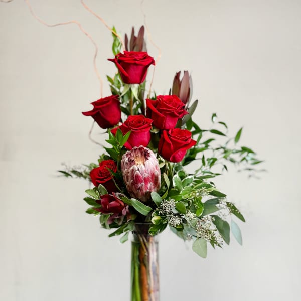Tall vase arrangement of red roses with greenery and a protea