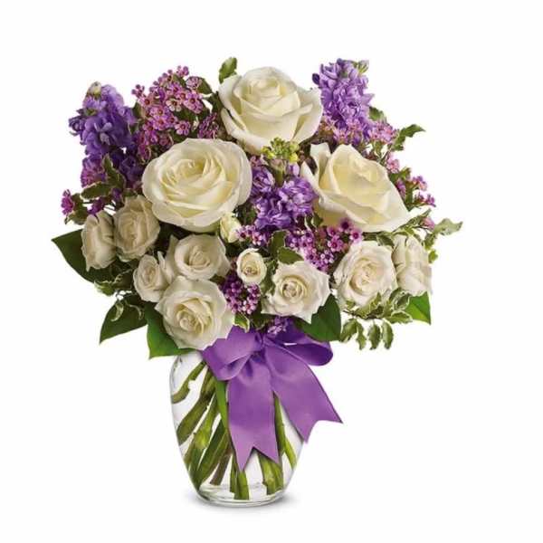 White roses and purple flowers arranged in a clear glass vase with a purple ribbon bow