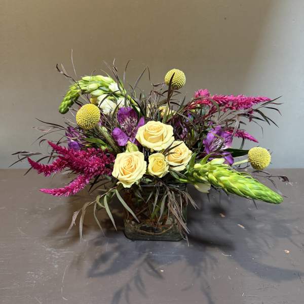 Mixed bouquet of yellow roses, purple flowers, and bright pink spikes in a glass vase