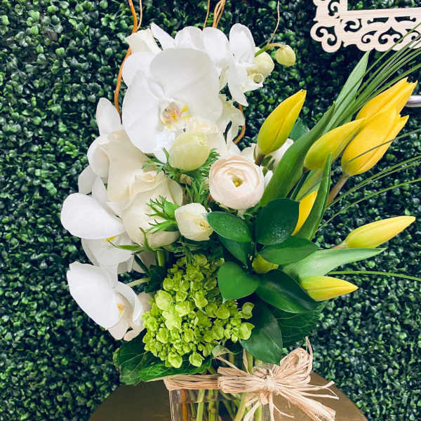 White orchids and yellow tulips in a glass vase with a raffia bow