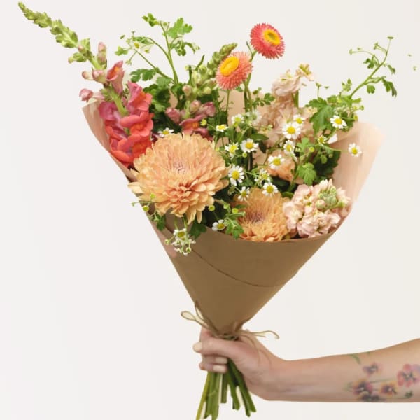 Handheld bouquet of peach, pink, and white mixed flowers wrapped in brown paper