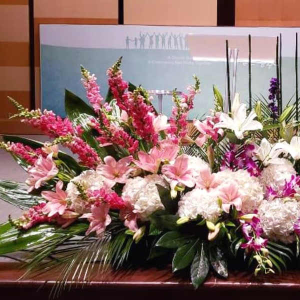 Large low centerpiece of pink lilies, white hydrangeas, and purple flowers along a stage edge