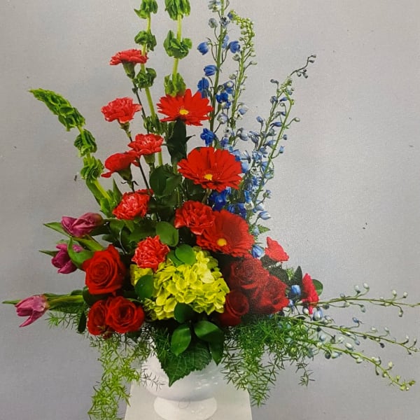 Tall mixed arrangement with red roses, gerbera, blue delphinium, and green hydrangea in a white vase