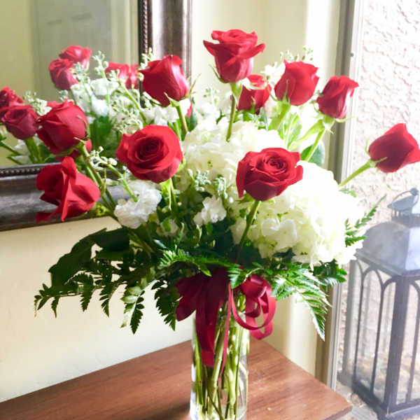 Tall arrangement of red roses and white flowers in a clear glass vase with red ribbon