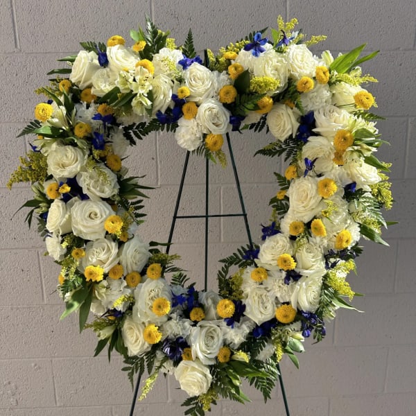 Heart-shaped floral wreath with white roses, yellow blooms, and blue accents on a stand