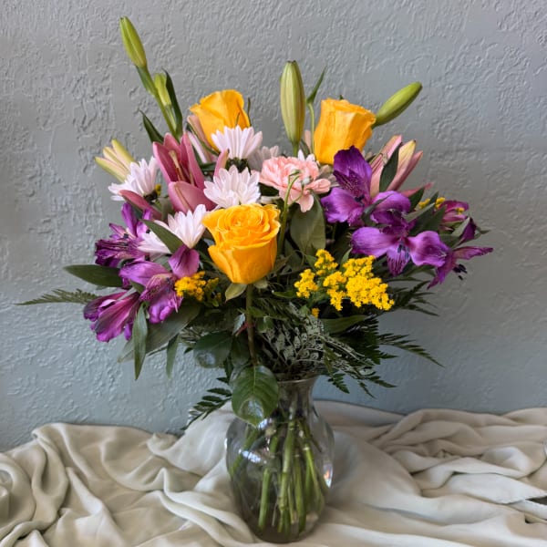 Mixed bouquet of yellow roses, purple alstroemeria, lilies, and daisies in a glass vase