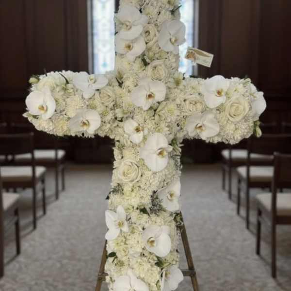Tall white floral cross with orchids and roses displayed on an easel in a chapel