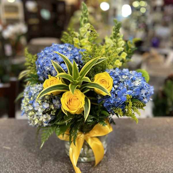 Blue hydrangeas and yellow roses in a glass vase with a yellow ribbon