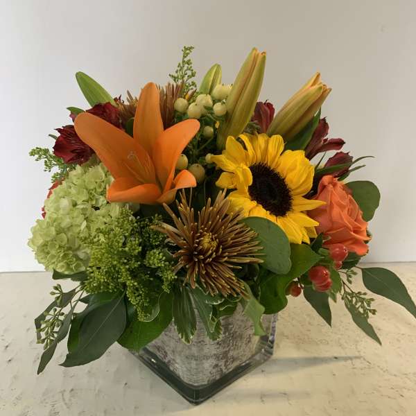 Mixed bouquet with orange lilies, a sunflower, and roses in a square glass vase