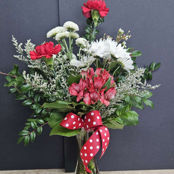 Bouquet of red carnations, white daisies, and pink alstroemeria in a glass vase with a red polka-dot ribbon.