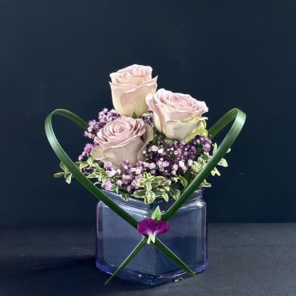 Three pale pink roses in a purple glass vase with green ribbon accents