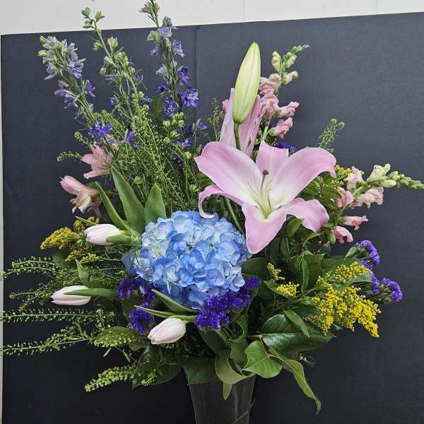 Tall mixed bouquet with pink lilies, blue hydrangea, and purple flowers in a black vase