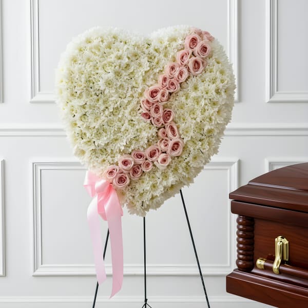 Heart-shaped white floral tribute with pink roses on a stand beside a casket
