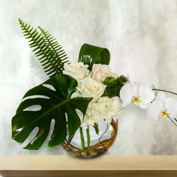 White roses and orchids arranged in a glass bowl with large tropical leaves