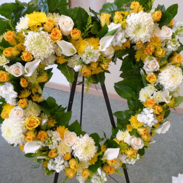 Heart-shaped standing wreath of yellow and white roses, mums, and calla lilies on a metal easel.