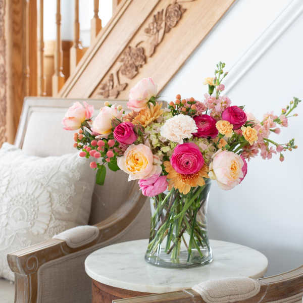 Mixed bouquet of pink, peach, and yellow flowers in a clear vase on a small table between chairs