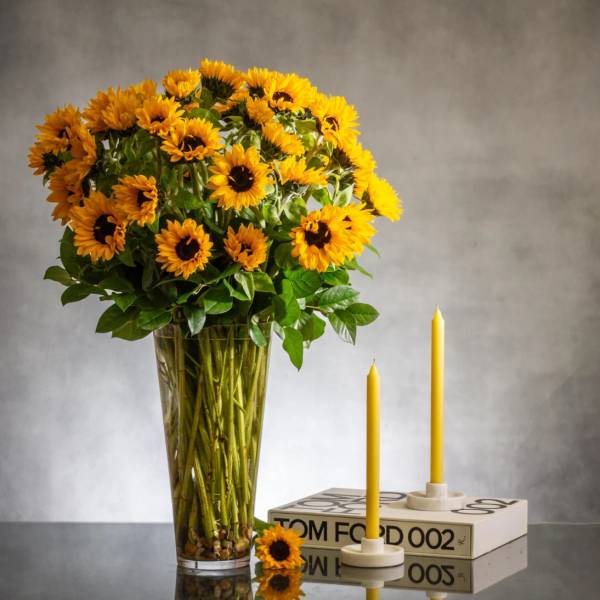 Tall arrangement of yellow sunflowers in a clear vase beside two yellow taper candles on a book.
