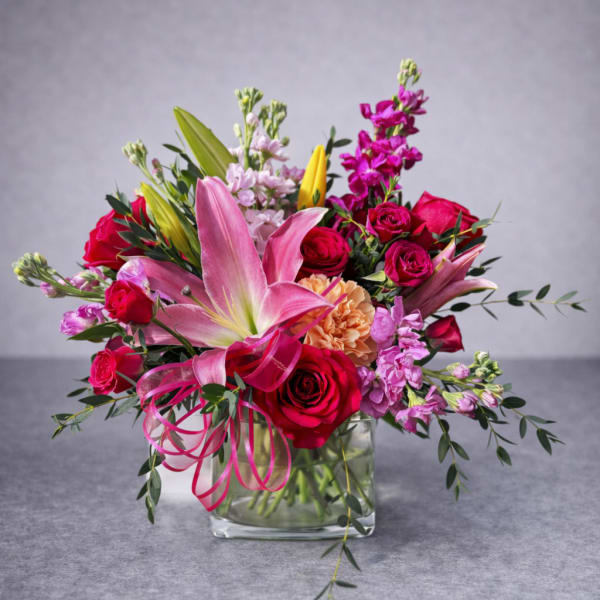 Pink lilies and red roses arranged in a clear glass vase with ribbon.