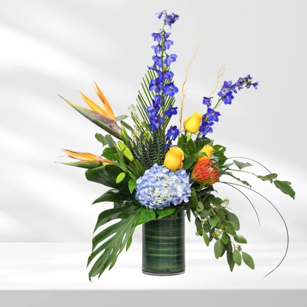 Tropical arrangement with blue flowers, yellow roses, and bird of paradise in a glass vase