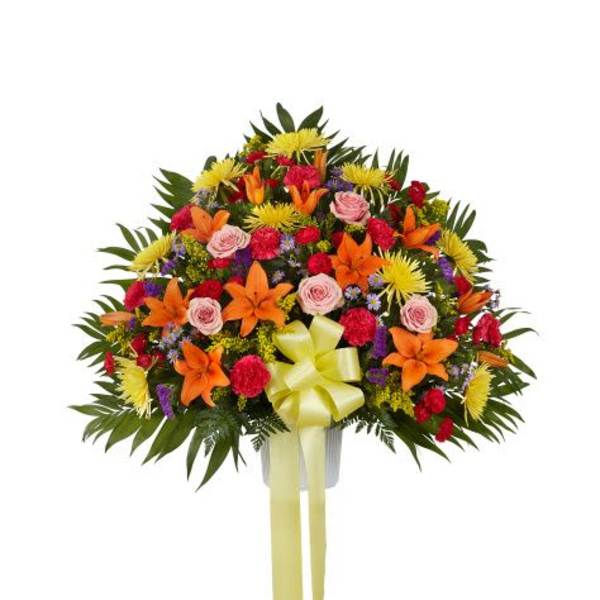 Fan-shaped arrangement of orange lilies, pink roses, red carnations and yellow mums in a white container with yellow bow
