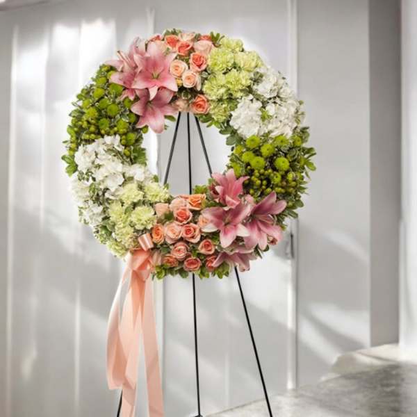 Floral wreath with pink lilies, peach roses, and white and green blooms on a black metal easel with peach ribbon