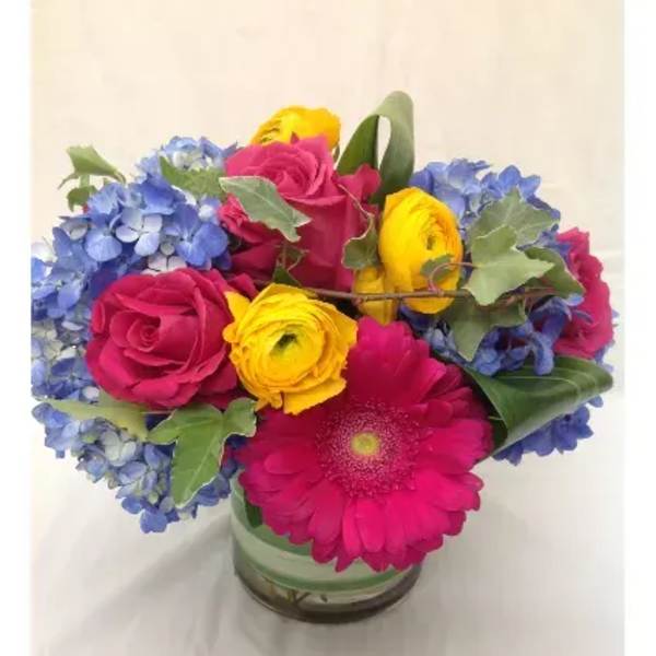 Bright bouquet of pink roses and gerbera daisy, yellow ranunculus, and blue hydrangeas in a glass vase