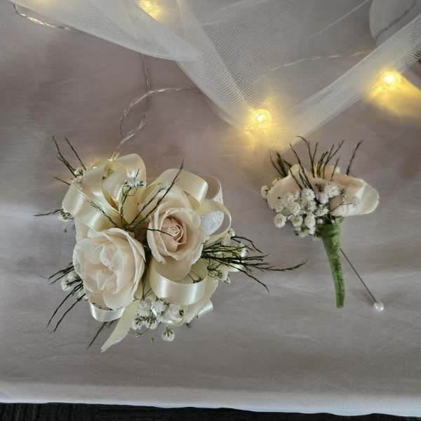 Ivory rose wrist corsage and matching boutonniere with ribbon on a draped cloth with soft lights.