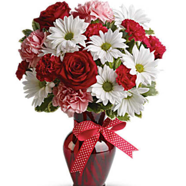 Bouquet of red roses, pink carnations, and white daisies in a red vase with a ribbon