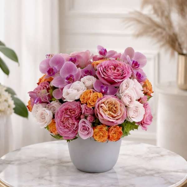 Low round arrangement of pink roses, orchids, and orange spray roses in a white pot on a marble table
