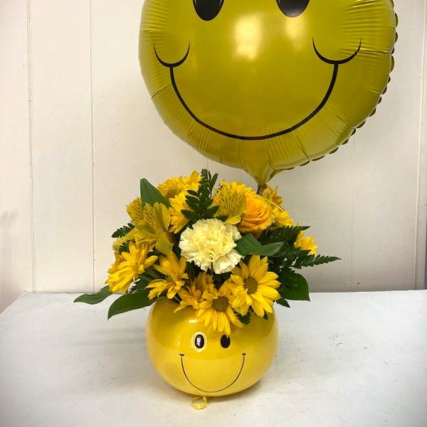 Yellow smiley-face balloon above a yellow flower arrangement in a matching container