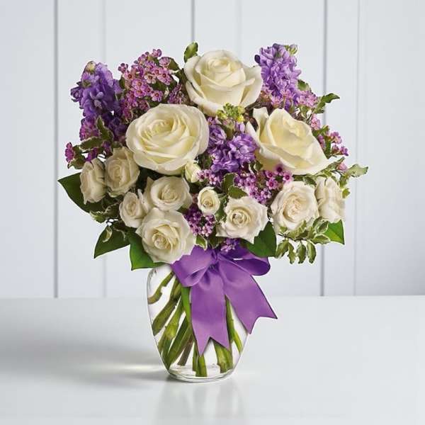 White roses and purple flowers in a clear glass vase with a purple ribbon