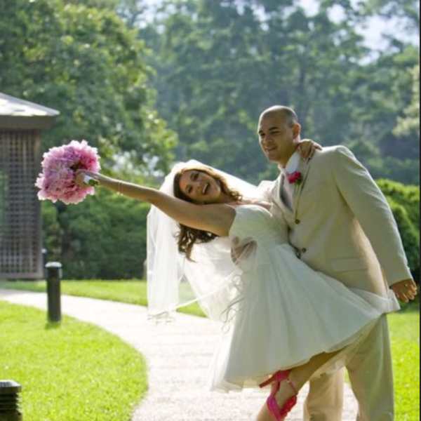 Bride being carried by groom, holding a pink bouquet outdoors