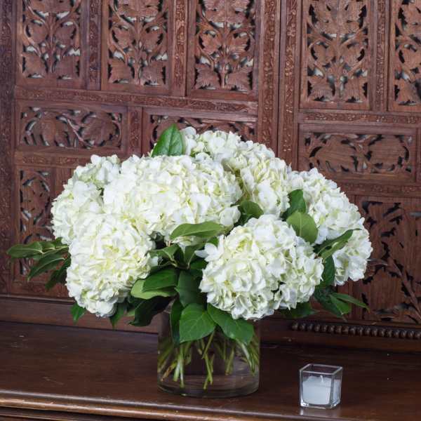 White hydrangea arrangement in a clear glass vase with a small candle beside it