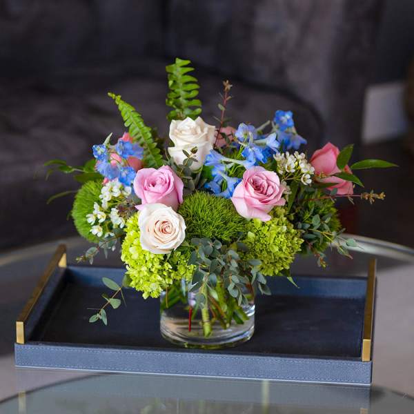 Low arrangement of pink and ivory roses with blue flowers in a clear glass vase
