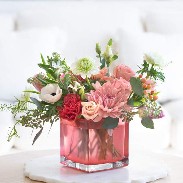Low pink and white mixed flower arrangement in a pink glass cube vase on a light table