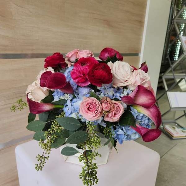 Low arrangement of pink roses, red ranunculus, blue hydrangeas, and dark pink calla lilies in a white vase