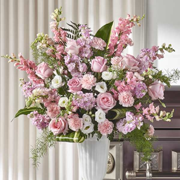 Tall arrangement of pink roses, carnations, and white flowers in a white urn on a pedestal