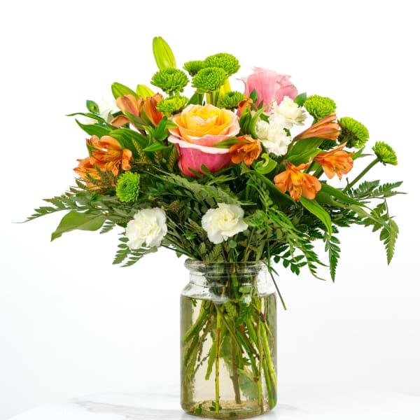 Bright mixed bouquet of roses, orange alstroemeria, green button mums, and white blooms in a clear glass jar.