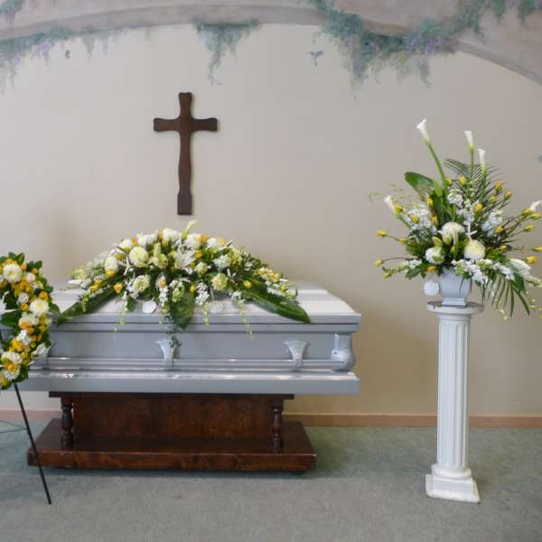 Funeral casket with white and yellow floral sprays, heart wreath, and pedestal arrangements.
