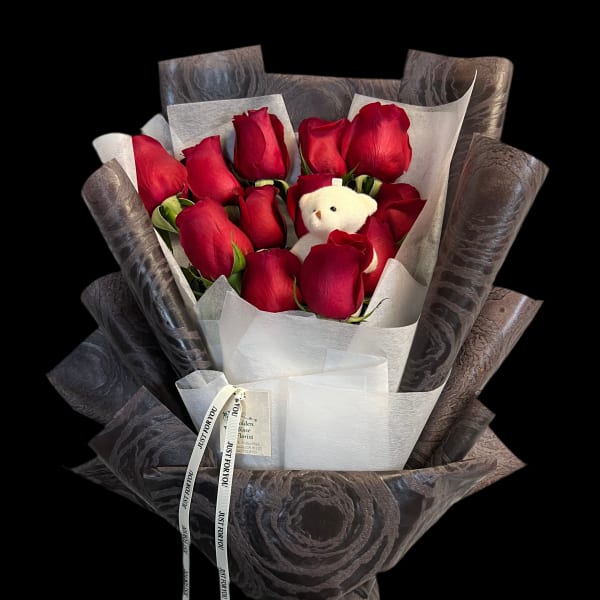 Hand-tied bouquet of red roses wrapped in dark paper with a small white teddy bear.