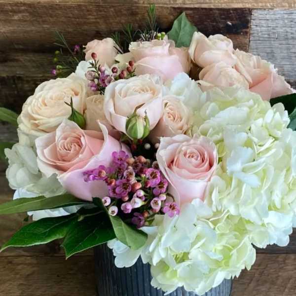 Short arrangement of pale pink roses, white hydrangeas, and small purple blooms in a gray vase