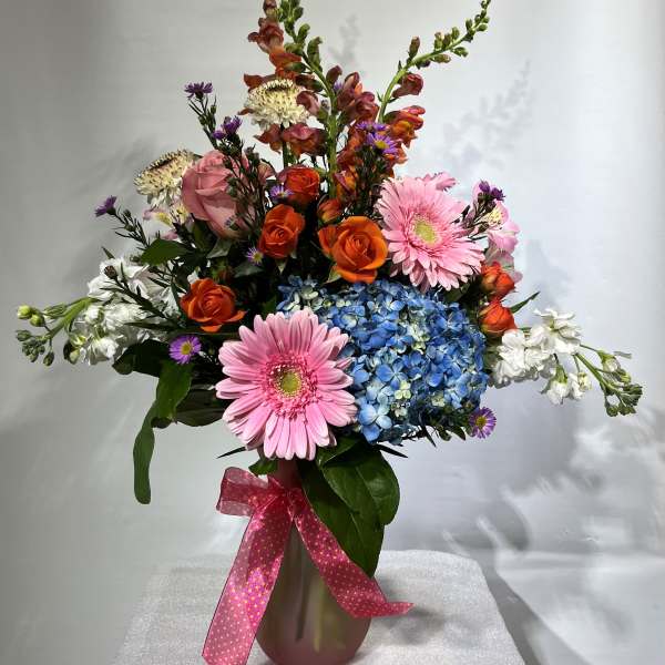 Mixed bouquet with pink gerberas, orange roses, blue hydrangea, and white blooms in a vase