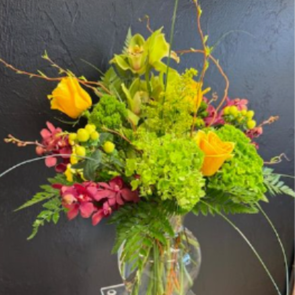 Tall arrangement of orange roses, green hydrangea, and orchids in a clear glass vase