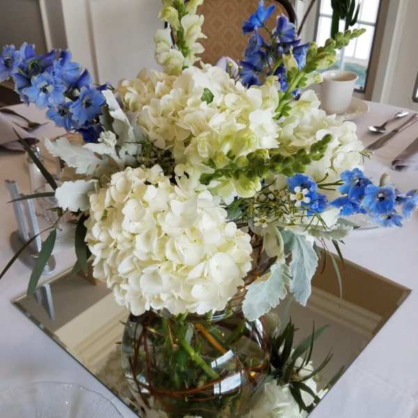 Centerpiece of white hydrangeas, snapdragons, and blue delphinium in a clear vase on a mirror.