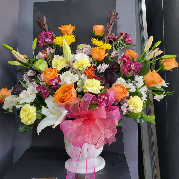 Mixed bouquet of orange roses, lilies, and carnations in a white vase with a pink bow