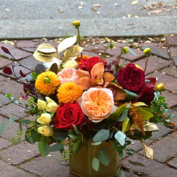 Autumn-tone arrangement of red roses, peach blooms and yellow flowers in a gold cube vase