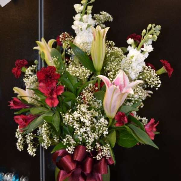 Tall bouquet of lilies, red flowers, and baby's breath in a vase with a ribbon