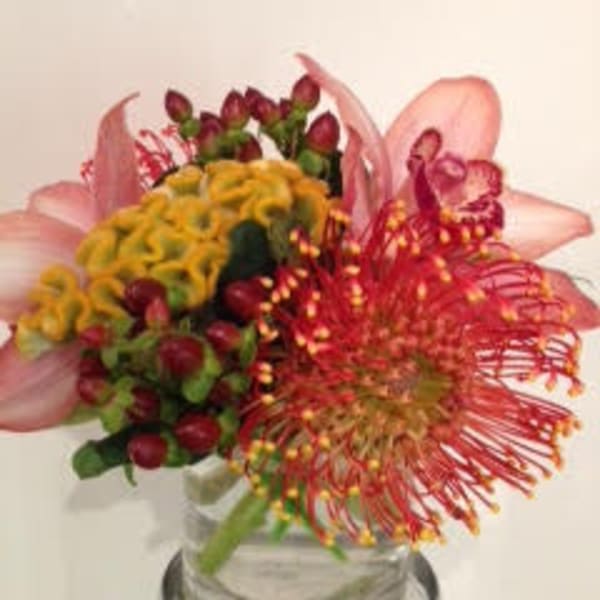Compact arrangement of pink lilies, red pincushion protea, yellow blooms, and berries in a clear glass vase.